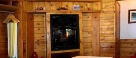 A wooden cabin interior with a bed, a shelf with various items, and a fireplace.
