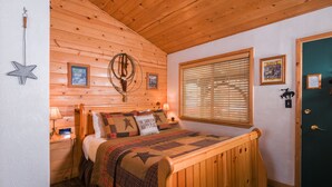 A cozy wooden cabin bedroom with a bed, a nightstand, a wall-mounted lamp, and a window with blinds. - Fireside Lodge (South Lake Tahoe)