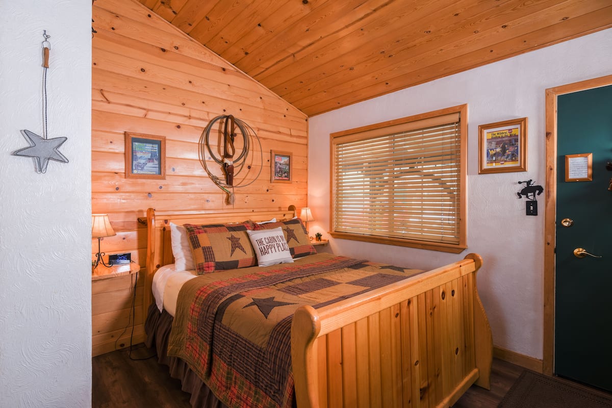 A cozy wooden cabin bedroom with a bed, a nightstand, a wall-mounted lamp, and a window with blinds.