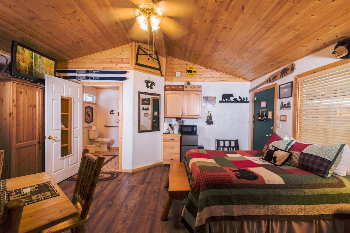 A cozy cabin-style room with a wooden ceiling, a bed with a quilt, a dining table with chairs, a bench, and a small kitchen area.