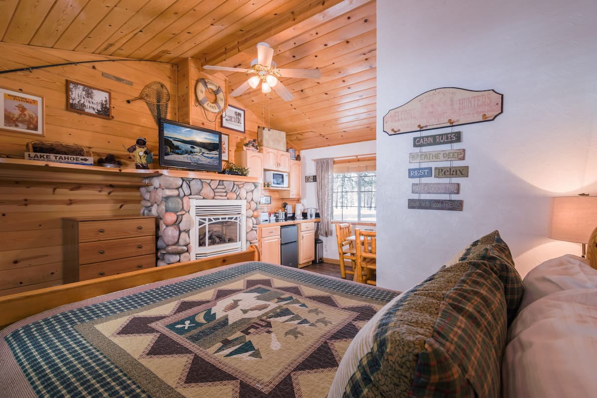 A cozy cabin interior with a wooden ceiling, a fireplace, a kitchen area, and a bed with a quilt.
