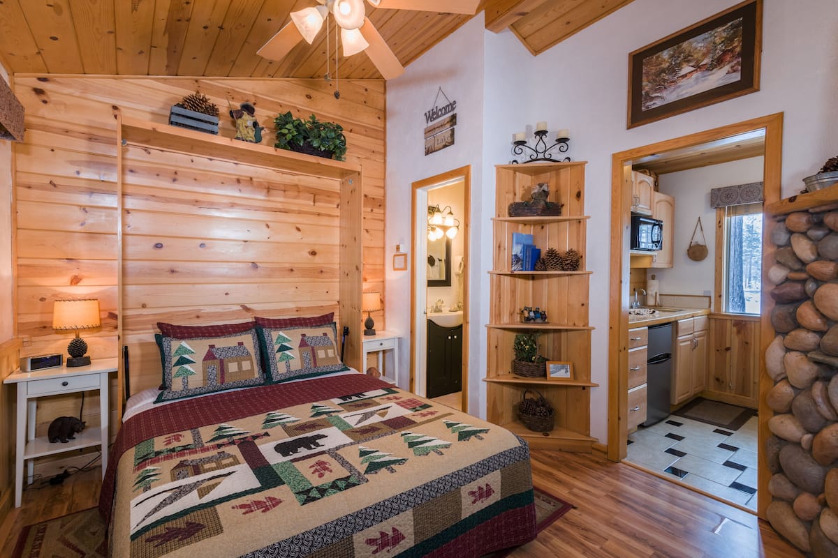 A cozy wooden cabin interior with a bed, a small desk, a kitchen area, and a stone wall.