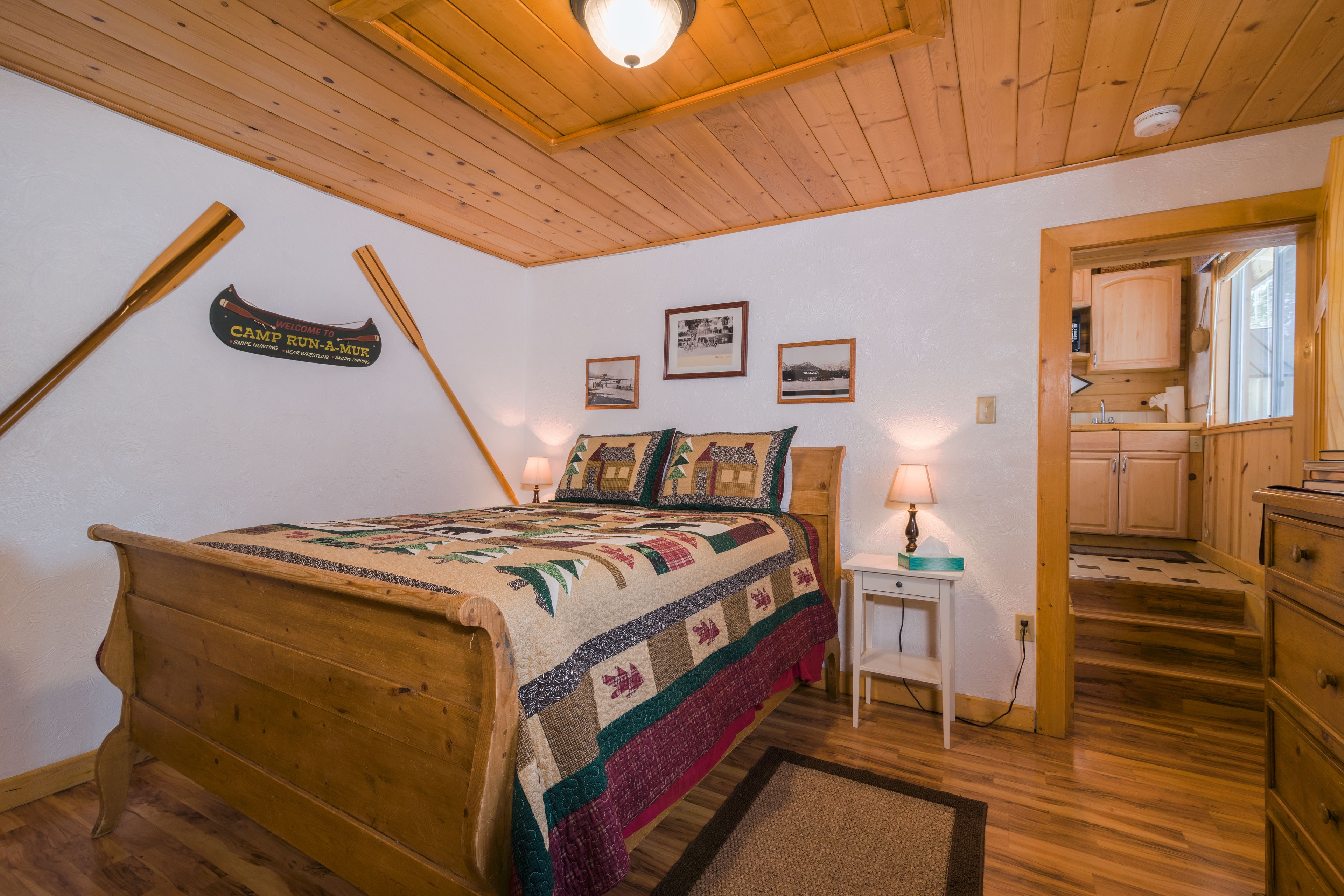 Une chambre confortable avec un lit en bois, une courtepointe, un panneau d’accueil, des photos encadrées et une petite table avec une lampe.
