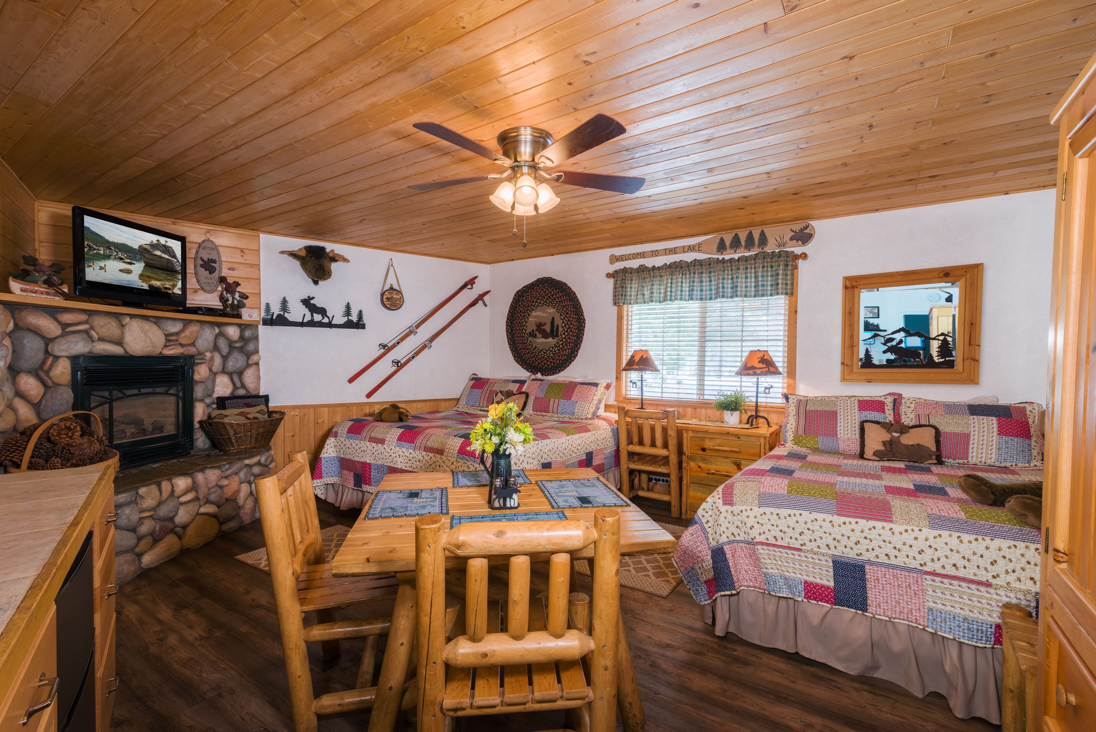 A cozy cabin interior with a stone fireplace, wooden furniture, a quilted bed, and a ceiling fan.
