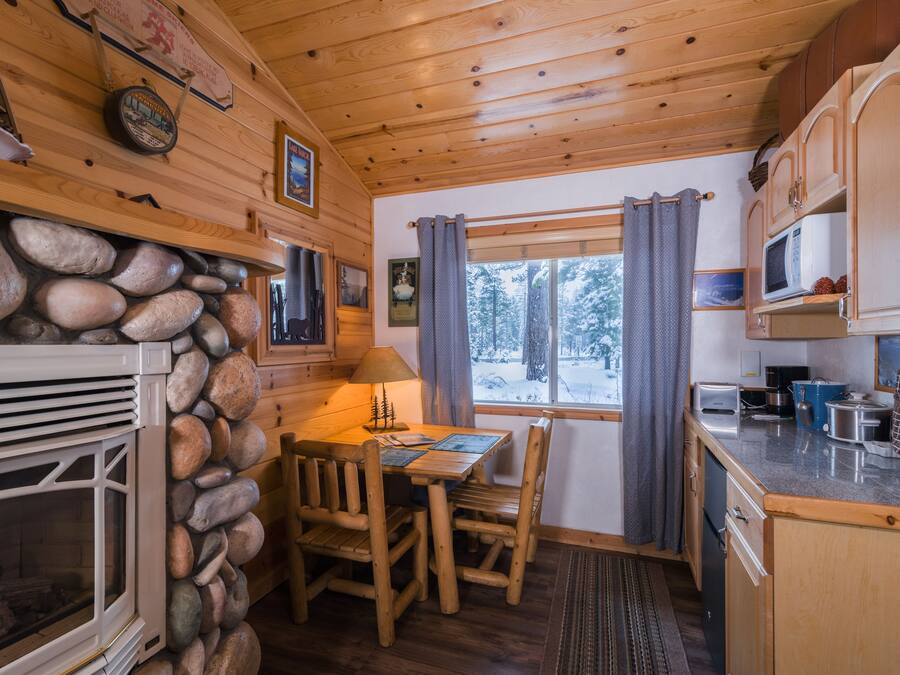 A cozy cabin interior with a stone fireplace, wooden dining table, and kitchen area.