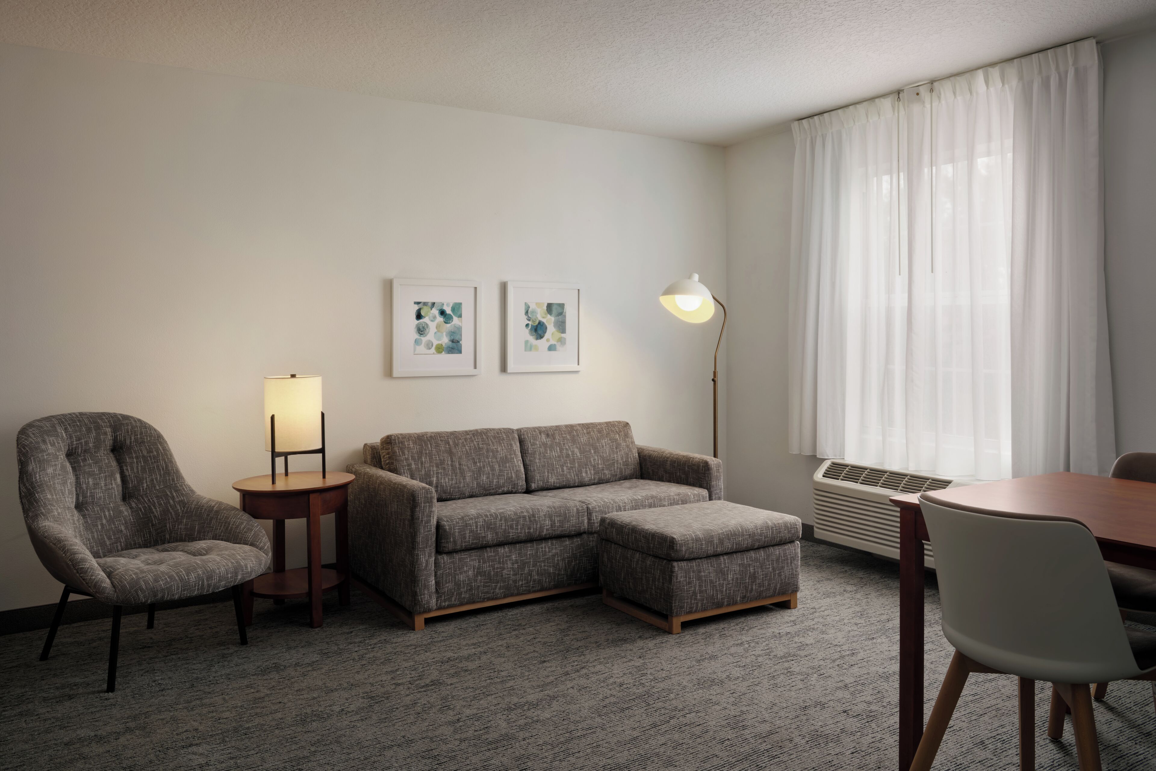 A hotel room with a grey sofa, a small armchair, a round table, and two wall-mounted lamps.