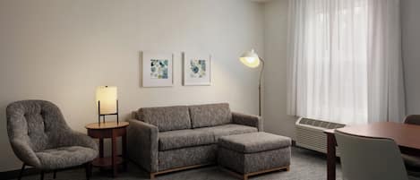 A hotel room with a grey sofa, a small armchair, a round table, and two wall-mounted lamps.