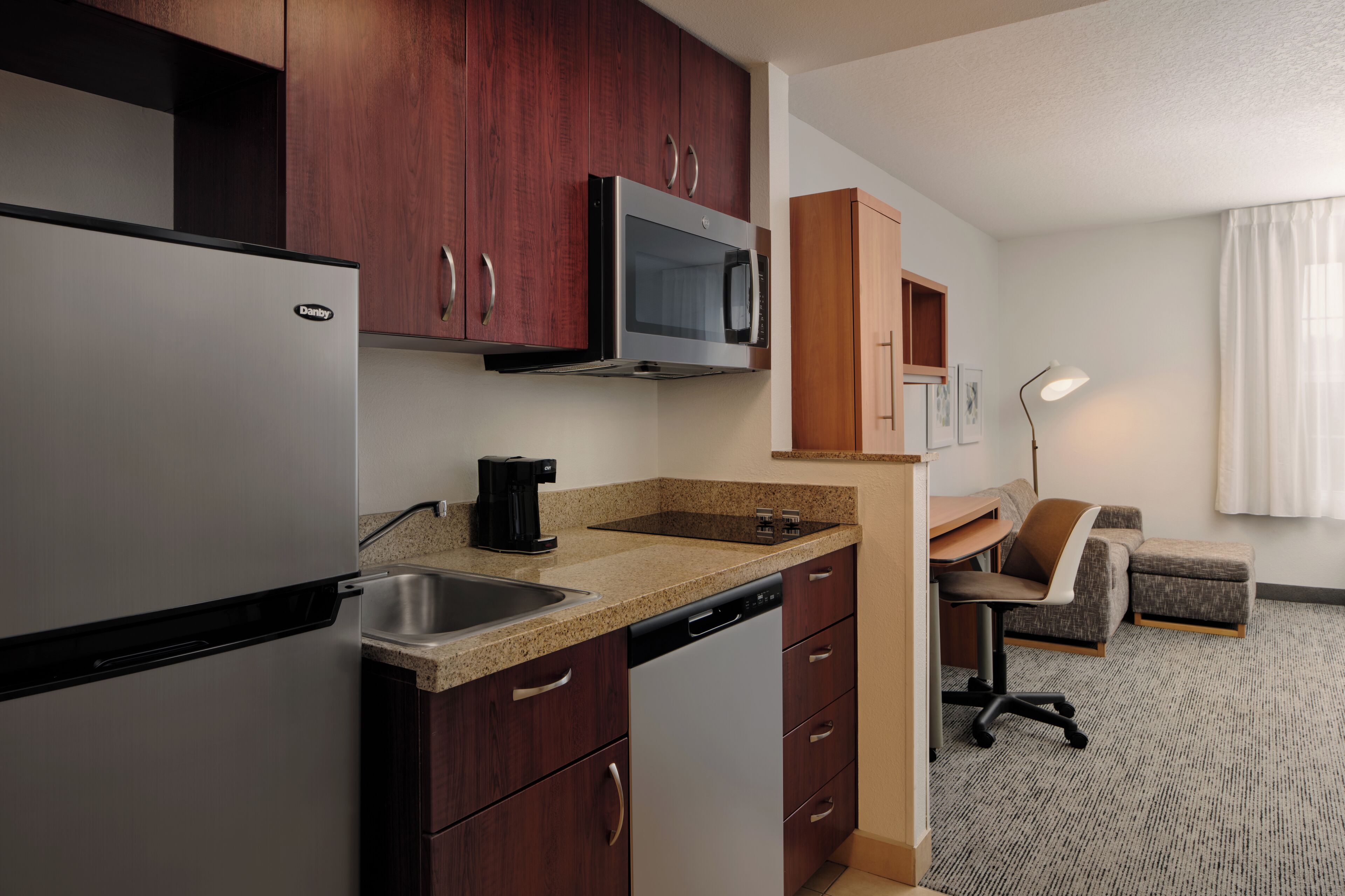 A compact kitchen with a refrigerator, microwave, coffee maker, and dishwasher. There is a small seating area with a desk and chair.