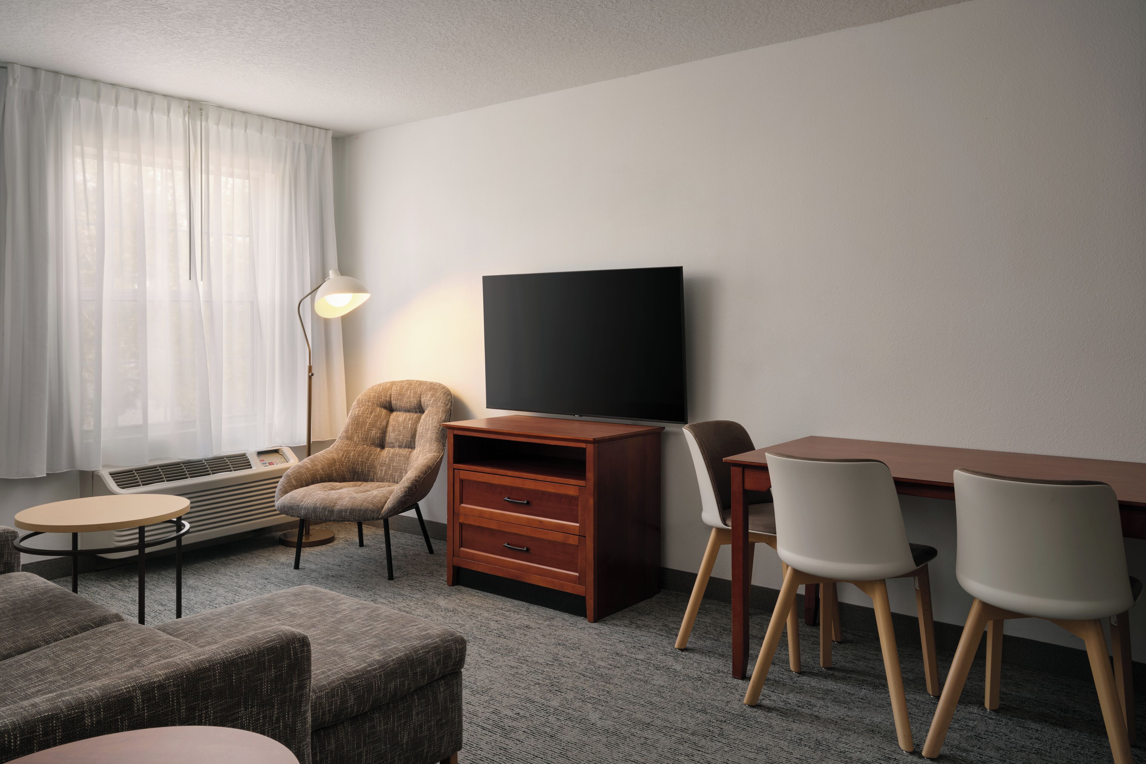 A hotel room with a flat-screen TV, a sofa, a chair, a small table, and a desk with chairs.