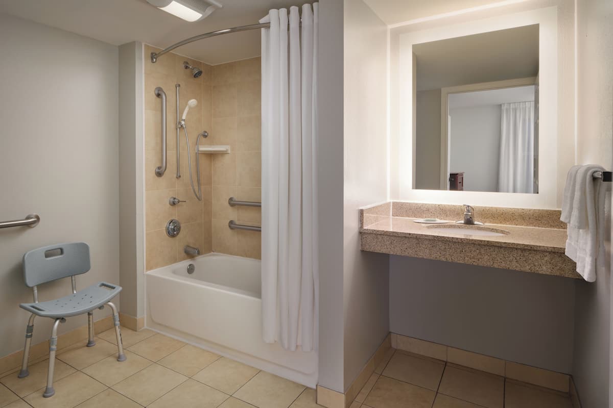 Une salle de bain équipée d’une douche, d’une baignoire, d’une barre d’appui, d’une poignée de maintien, d’un fauteuil roulant et d’un lavabo avec un miroir.