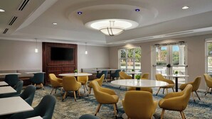 Un salón de conferencias de hotel con mesas redondas, sillas amarillas, un televisor de pantalla plana y ventanas amplias.