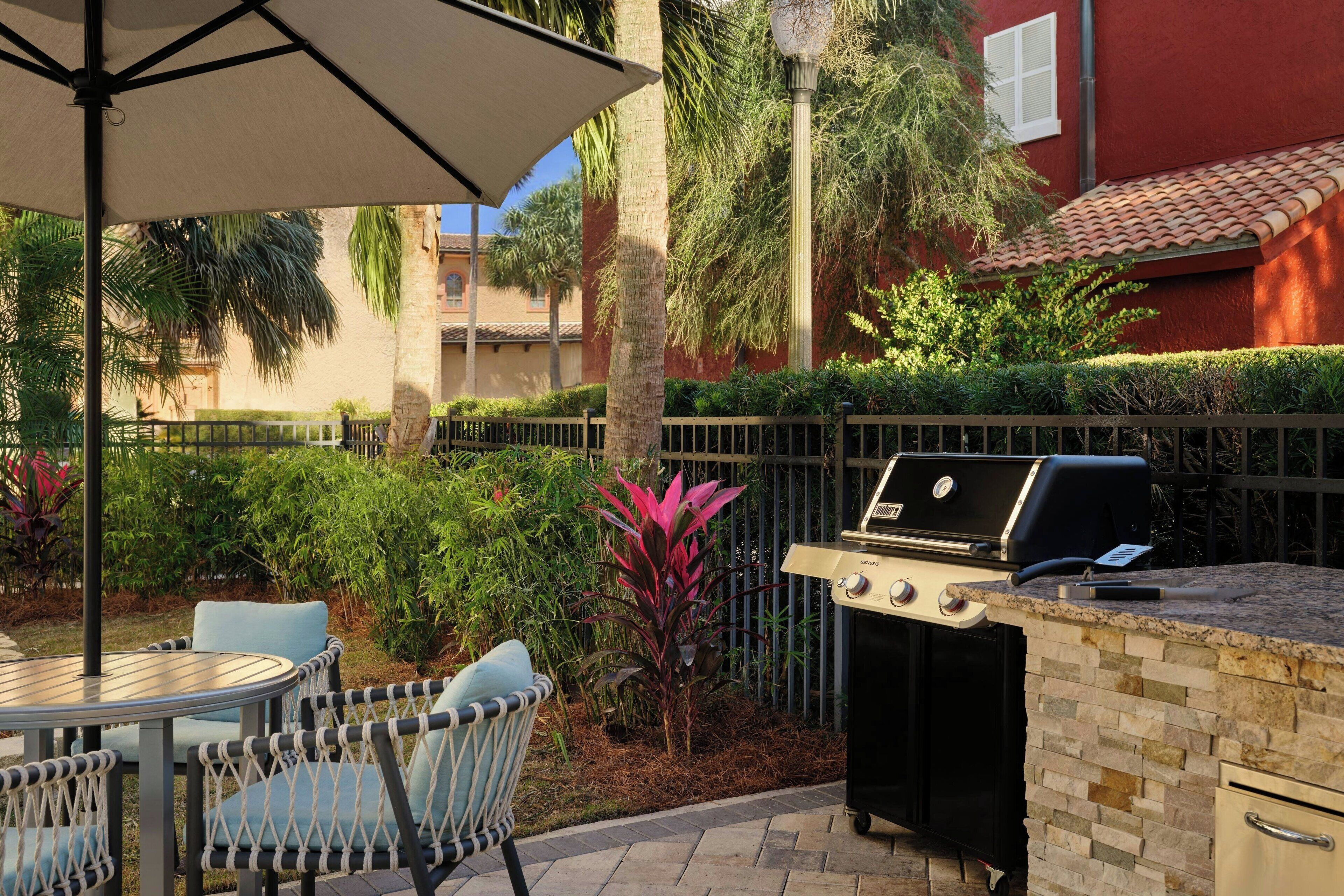 An outdoor patio with a grill, chairs, and a table under an umbrella.