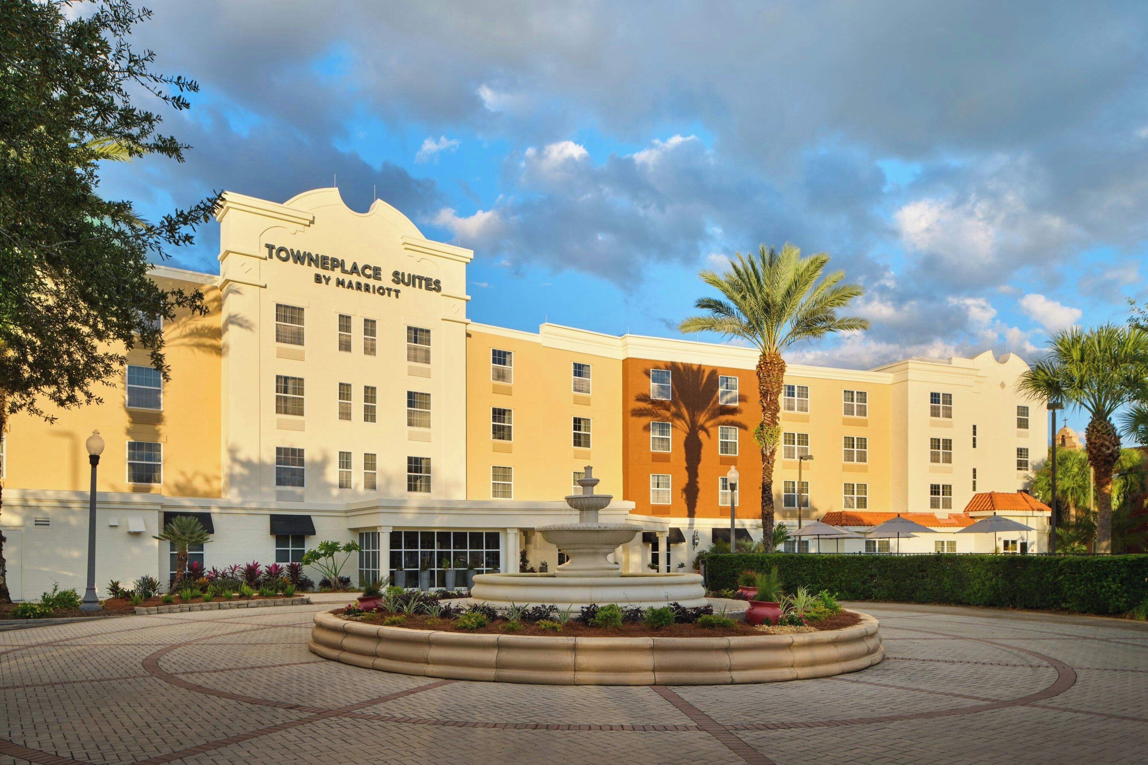 A hotel with a fountain in the courtyard, surrounded by palm trees and a paved walkway.