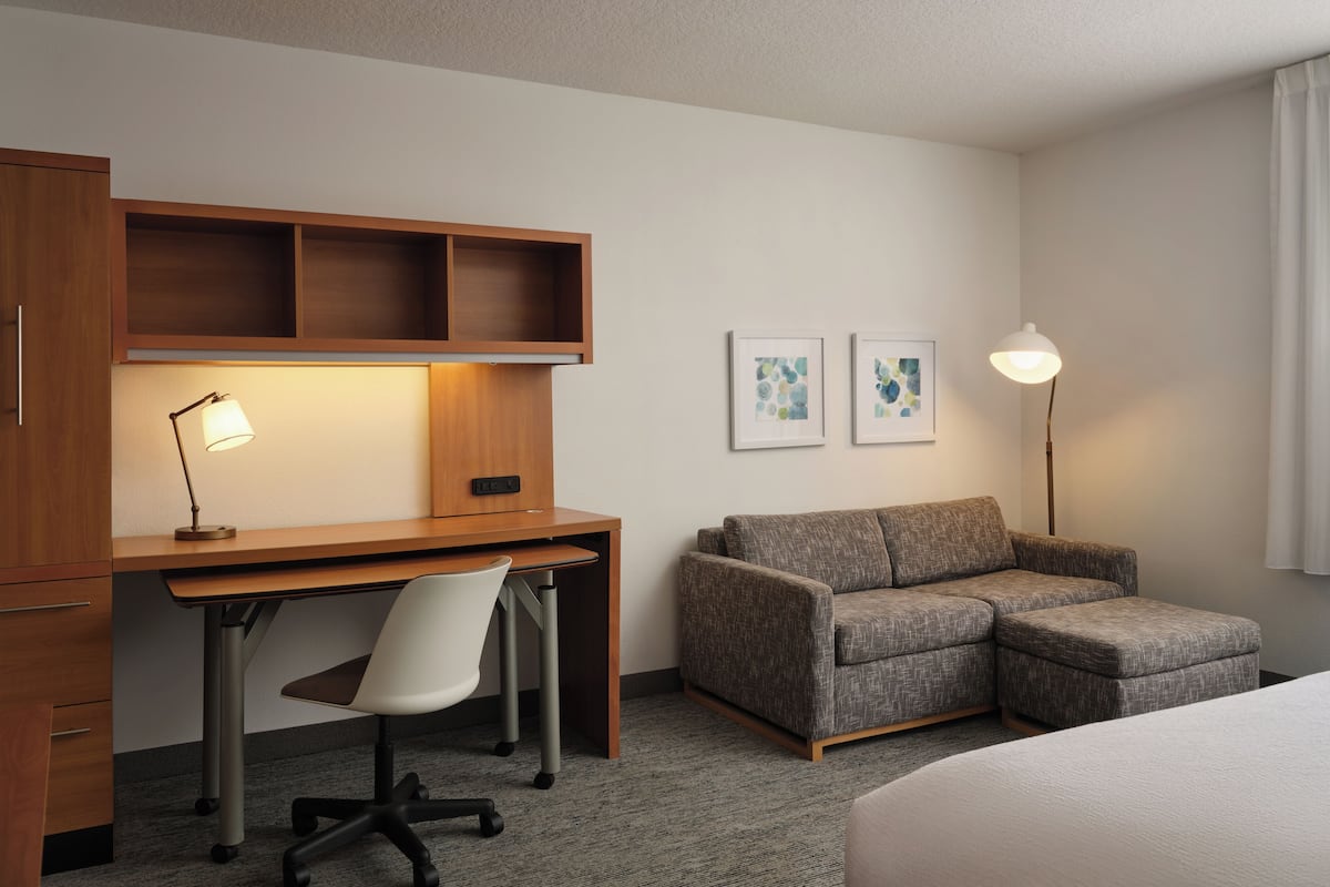Une chambre d’hôtel avec un bureau en bois, un canapé gris, une chaise blanche, un lit et deux œuvres d’art encadrées accrochées au mur.