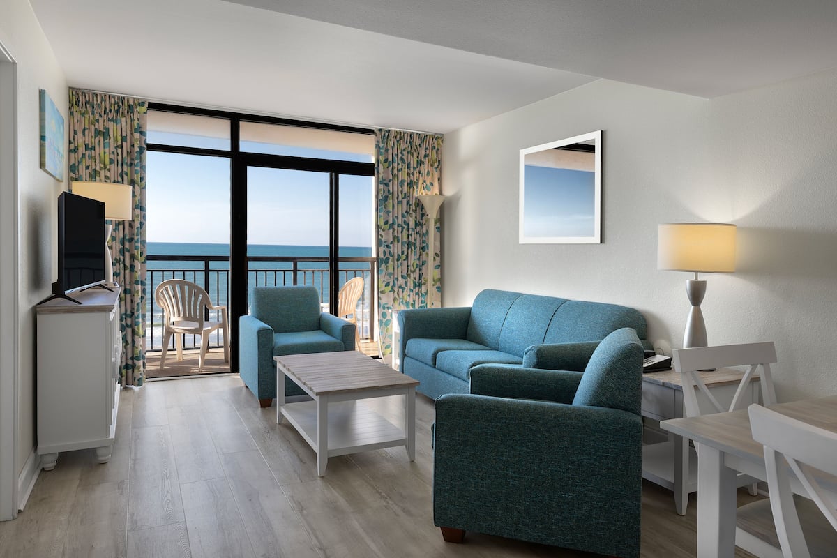 Quarto de hotel com varanda, sofá, poltrona, mesa pequena e televisão.