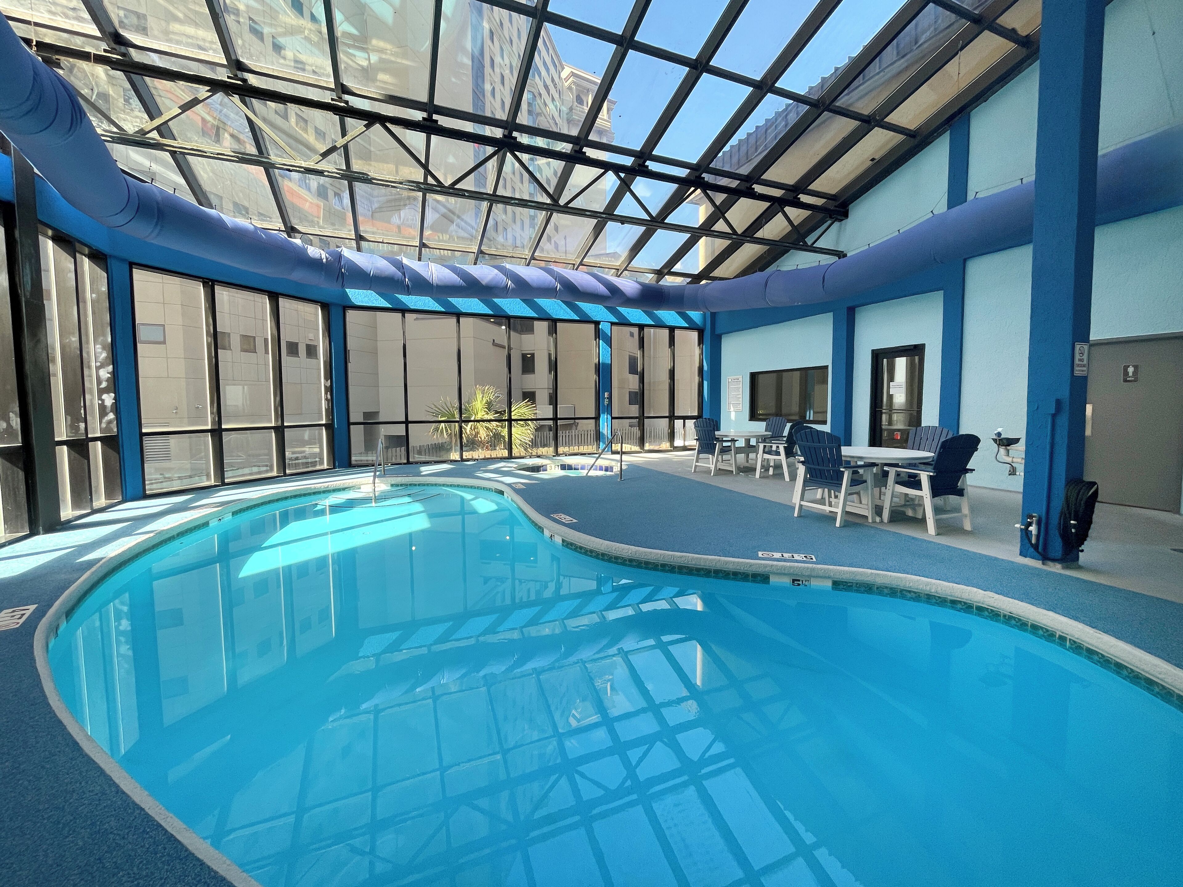 An indoor swimming pool with clear blue water, surrounded by blue walls and a glass ceiling. There are chairs and tables around the pool area.