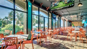 En rymlig restaurang med stora fönster, orangefärgade stolar och utsikt över palmer.
