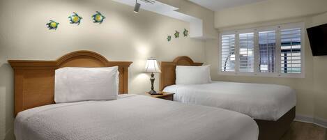A hotel room with two beds, a nightstand with a lamp, a ceiling light, wall art, and a window with blinds.