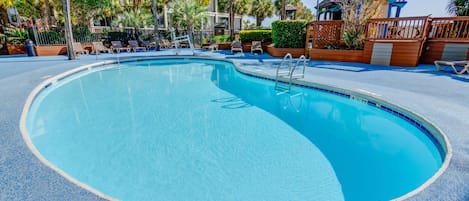 A large, clear swimming pool with a blue tiled edge, surrounded by lounge chairs and palm trees.