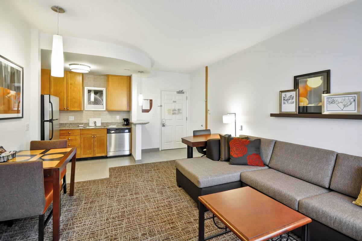 A modern living room with a sofa, coffee table, and dining area. There is a kitchen in the background with wooden cabinets and stainless steel appliances.