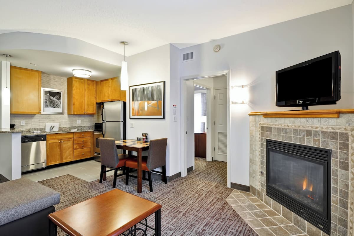 A modern kitchen with wooden cabinets, a dining area with a table and chairs, a flat-screen TV mounted above a fireplace, and a hallway leading to another room.