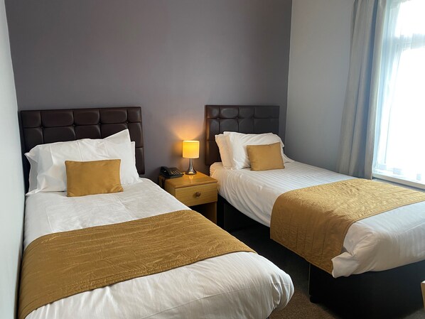Habitación de hotel con dos camas, cada una con cabecera, una mesita de noche con lámpara y una ventana con cortinas.