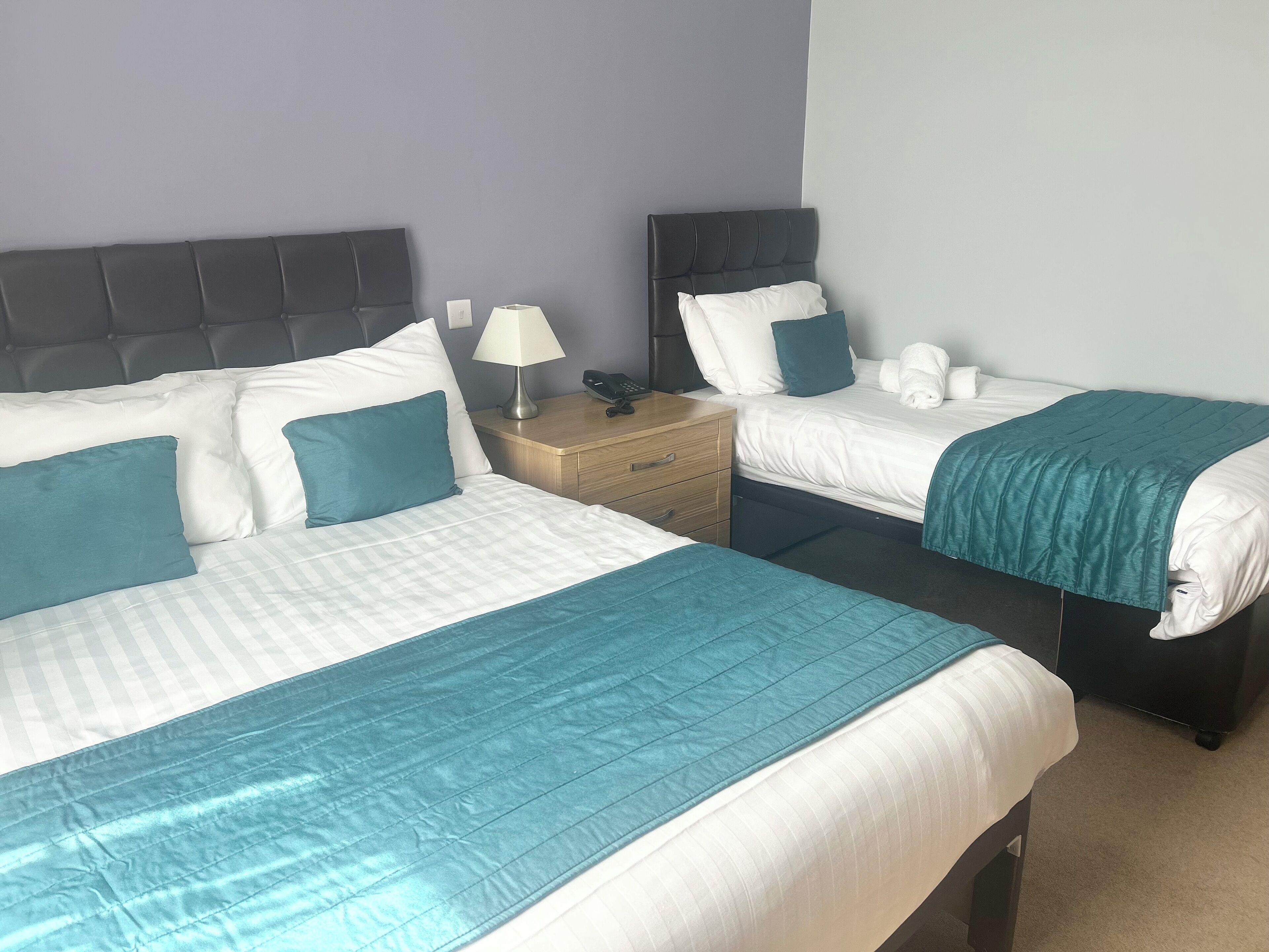Una habitación de hotel con dos camas, cada una con cubrecama color aguamarina y ropa de cama blanca. Entre las camas hay una mesita de noche de madera con una lámpara y un teléfono.