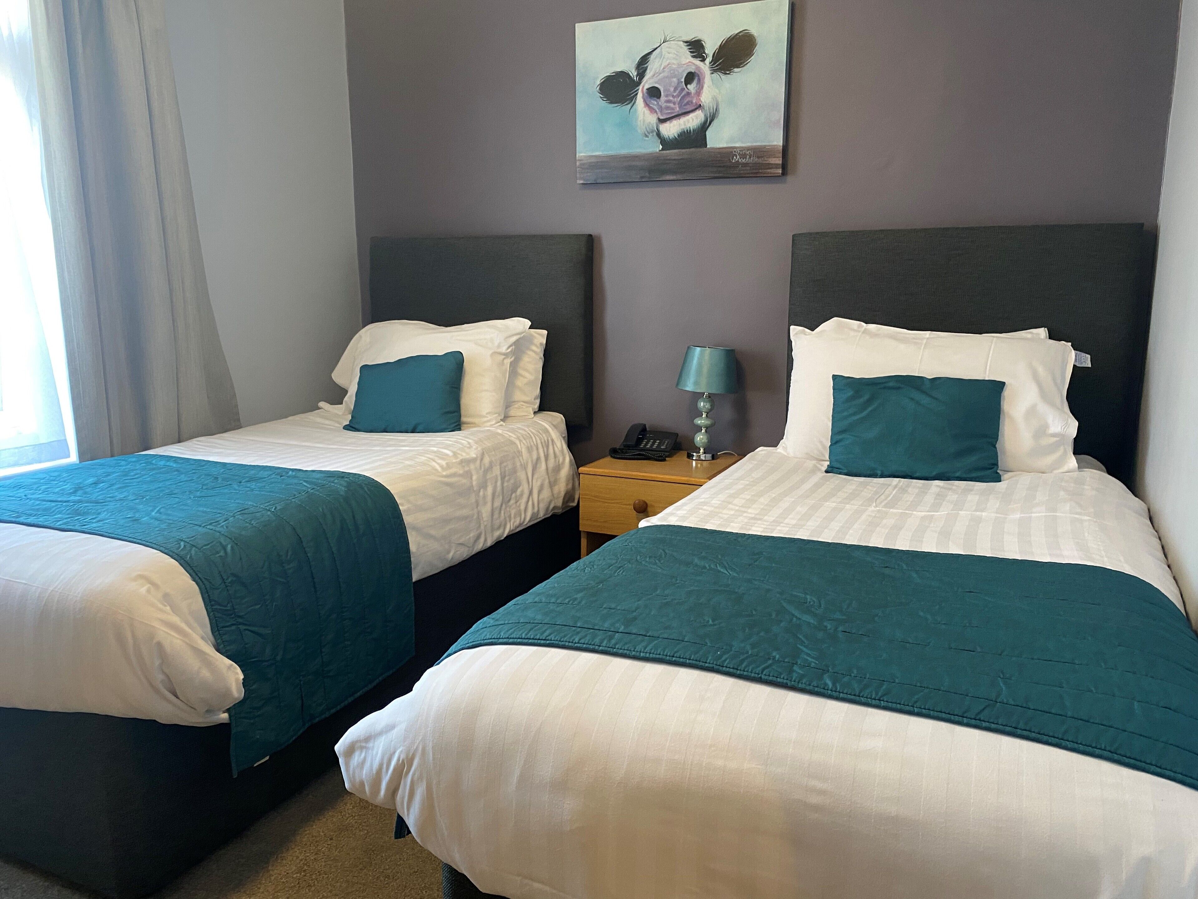 Two twin beds with teal bedding, a nightstand with a lamp, and a painting of a cow on the wall.