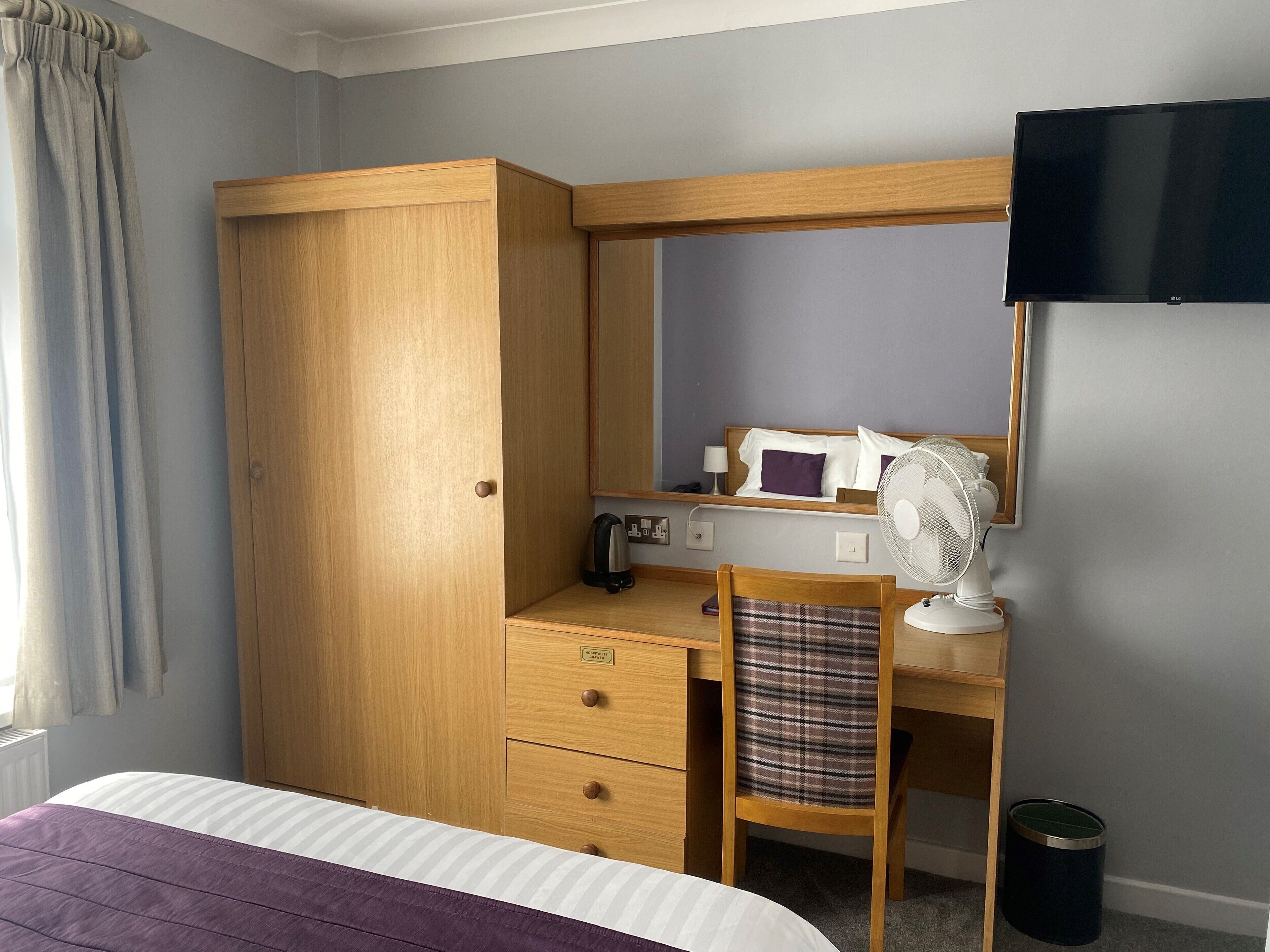 A bedroom with a bed, a wooden desk, a chair, a TV, and a small trash bin.