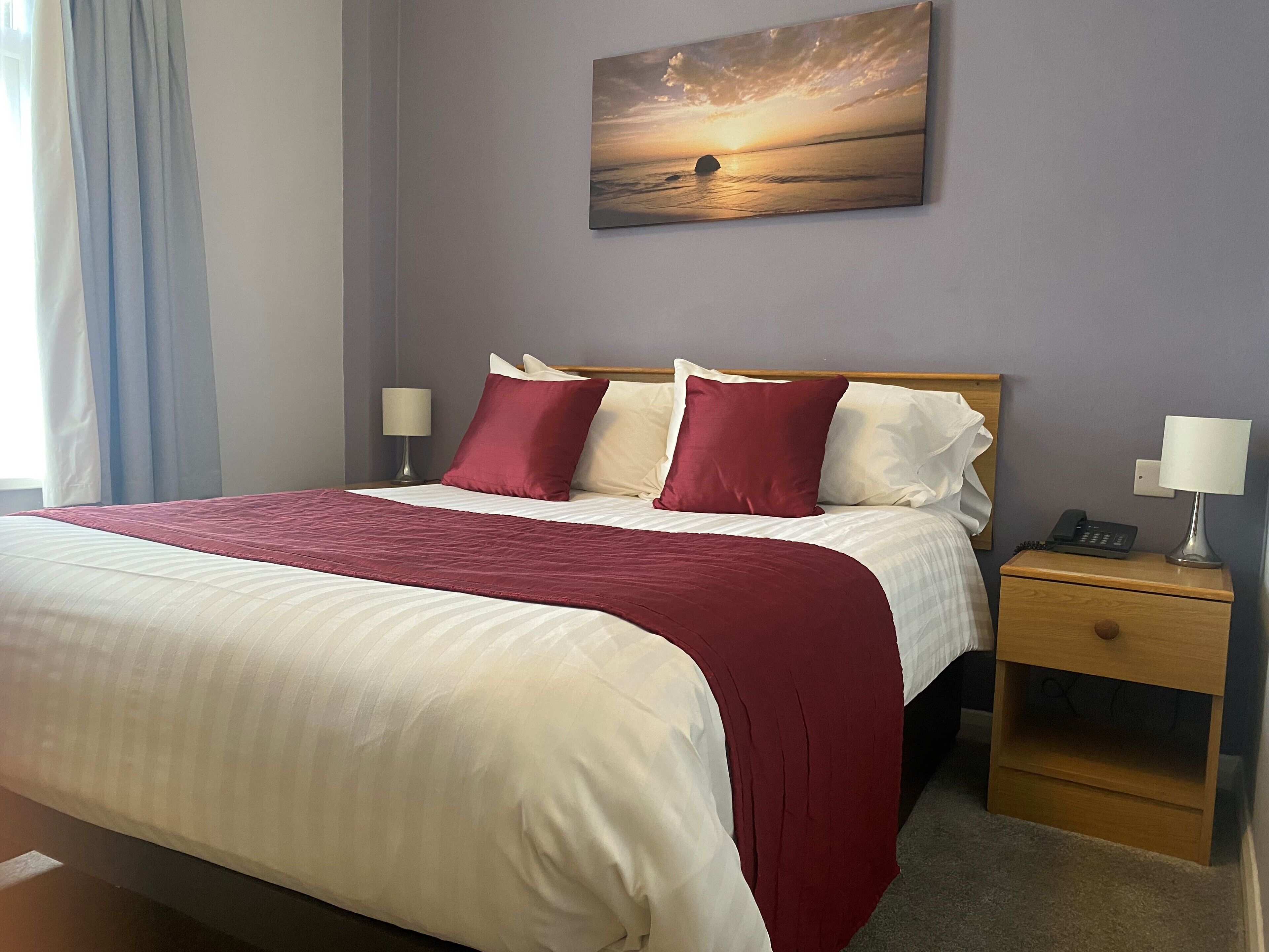 A neatly made bed with a red and white bedspread, two pillows, and a nightstand with a lamp and phone.