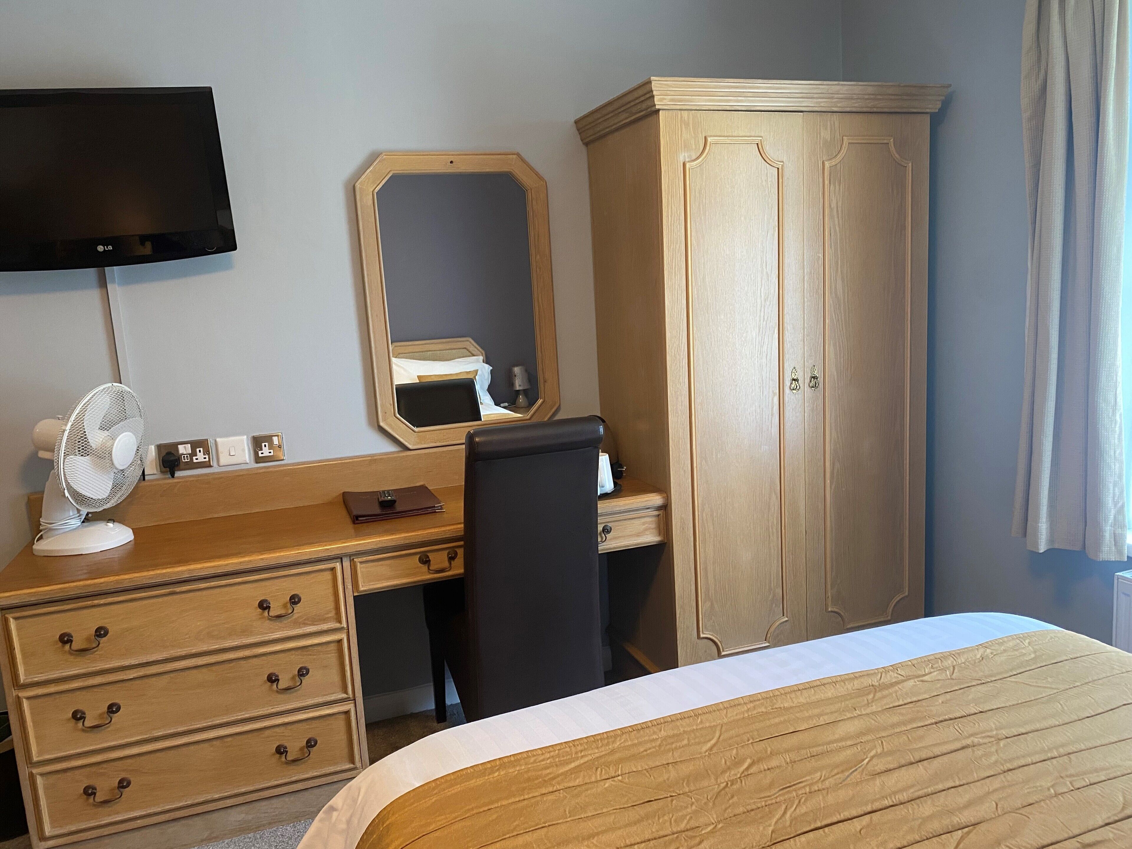 A hotel room with a bed, a desk with drawers, a chair, a wardrobe, a mirror, a flat-screen TV, and a standing fan.