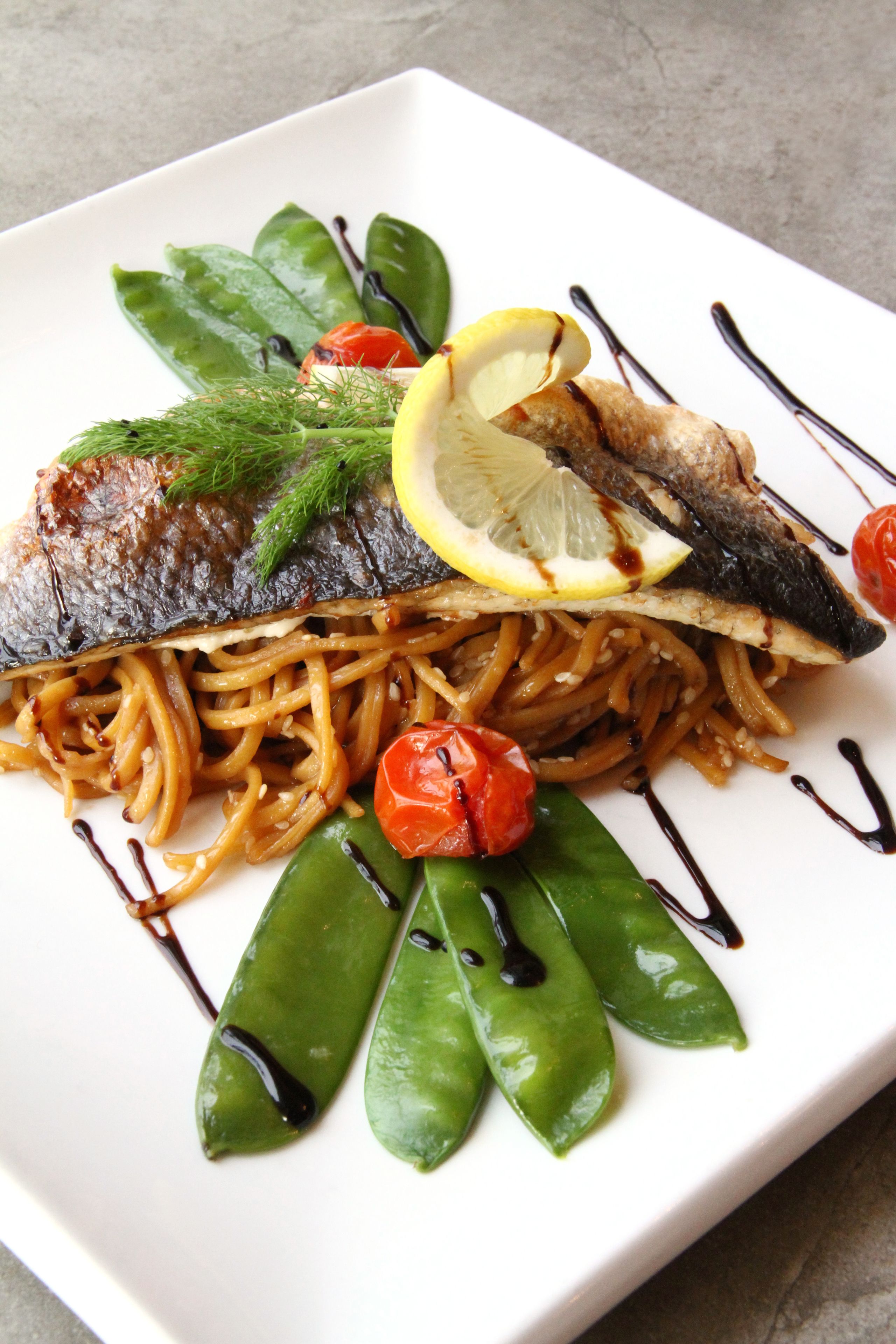 A square plate with a fish fillet, noodles, green vegetables, and a lemon slice.