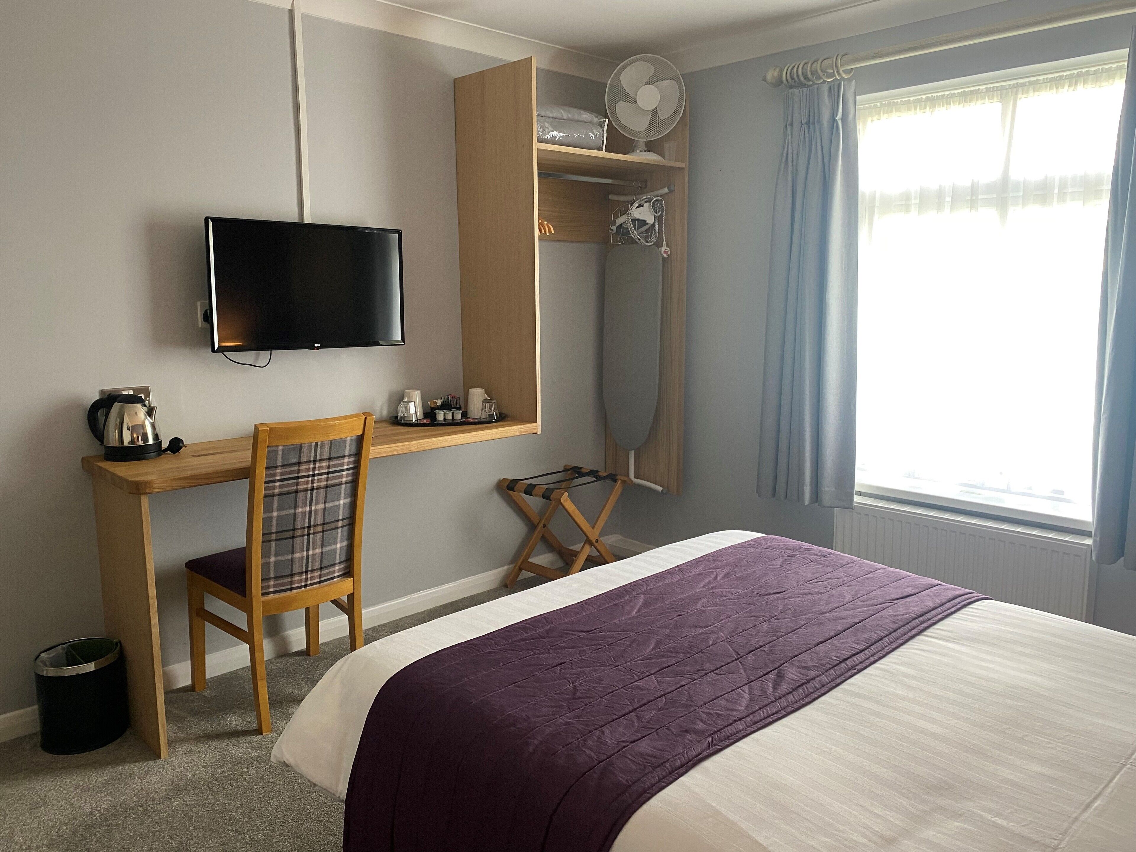 A hotel room with a bed, a desk with a chair, a television, a window with curtains, and a radiator.