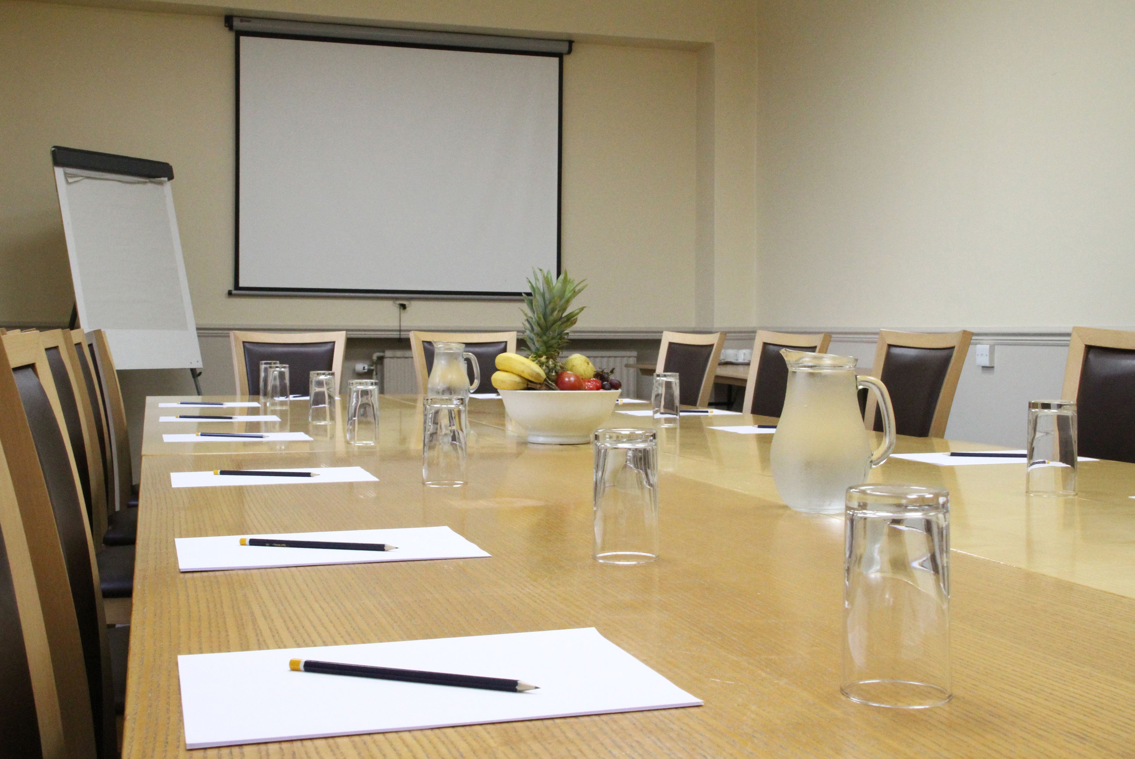 A conference room with a long table, chairs, a projector screen, a fruit bowl, and water glasses.
