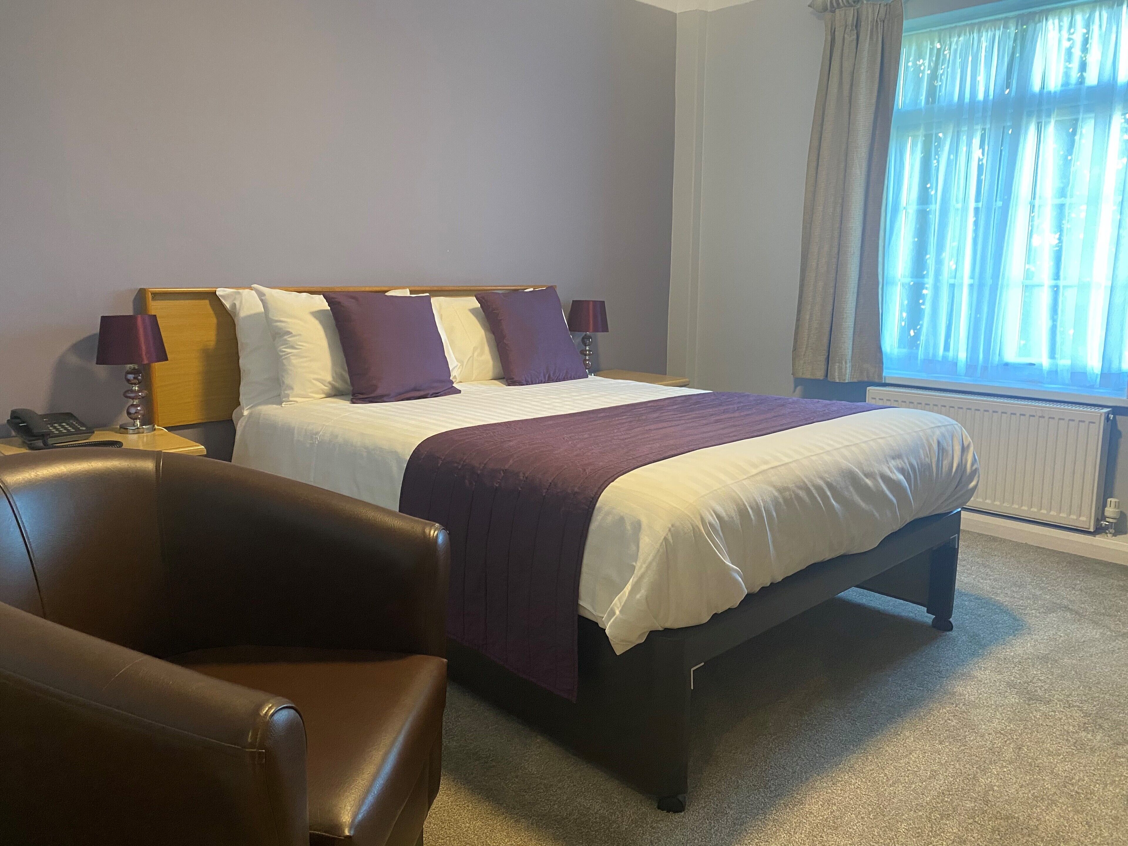 Una habitación de hotel con una cama, una silla, una mesita de noche con lámpara y una ventana con cortinas.