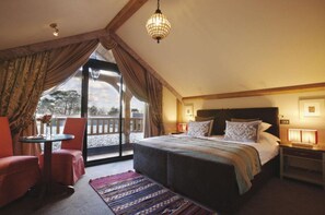 A bedroom with a large bed, a nightstand, a table with chairs, and a view of the outdoors.