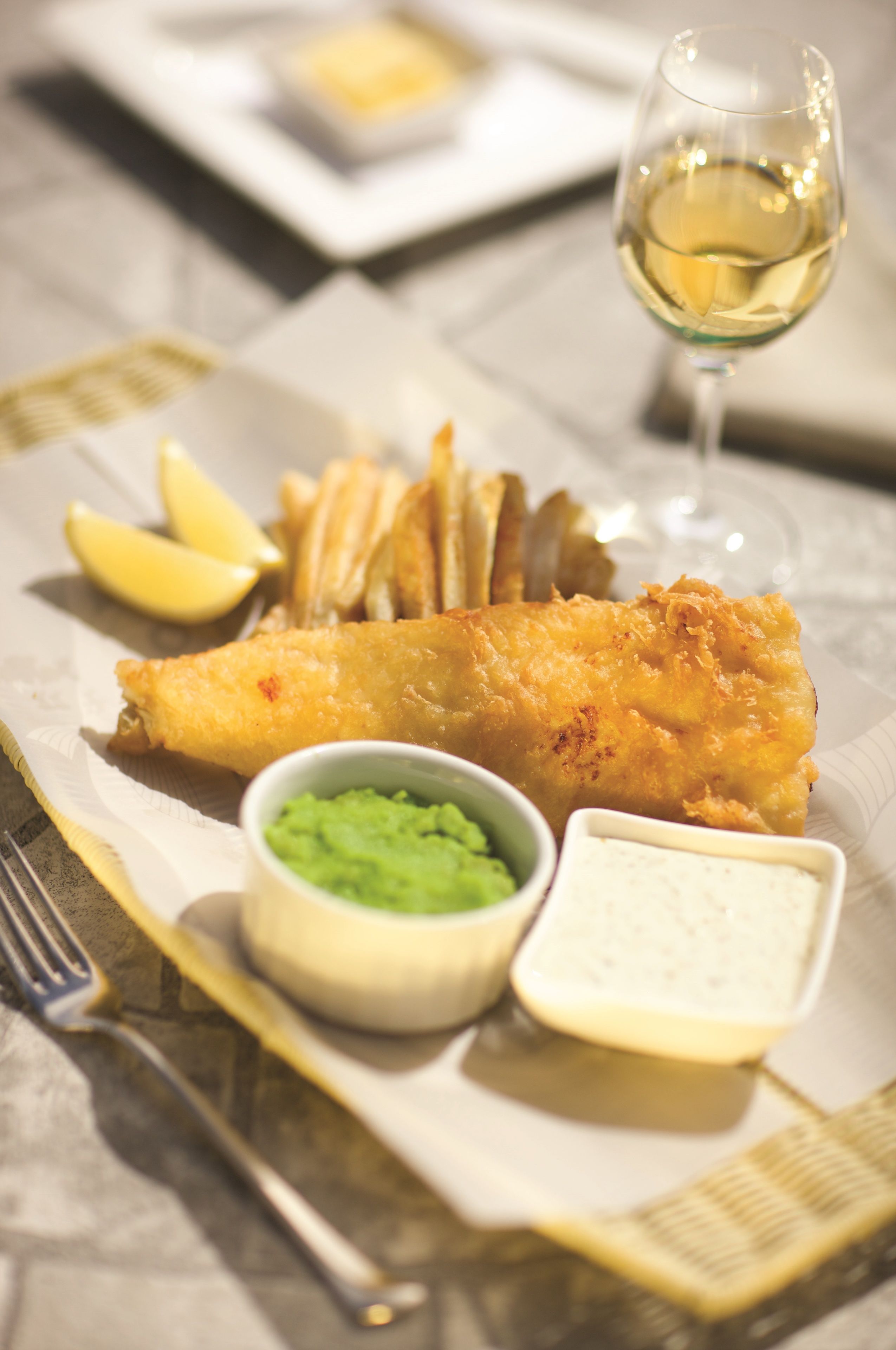A plate of fish and chips with lemon wedges, a glass of white wine, and two small bowls of sauce.
