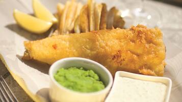 A plate of fish and chips with lemon wedges, a glass of white wine, and two small bowls of sauce.