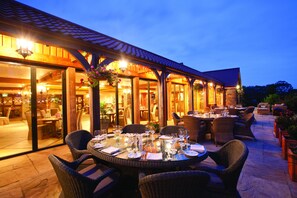 An outdoor dining area with wicker chairs and tables set for a meal, large windows, and a covered patio.
