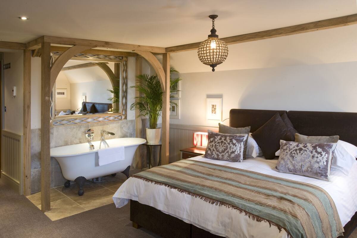 Quarto com uma cama grande, uma porta em arco de madeira que dá para um banheiro, um lustre e uma mesa de cabeceira com uma luminária.