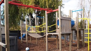 Een speeltuin met verschillende speeltoestellen, zoals een glijbaan en klimmogelijkheden, omringd door houtsnippers en bomen.