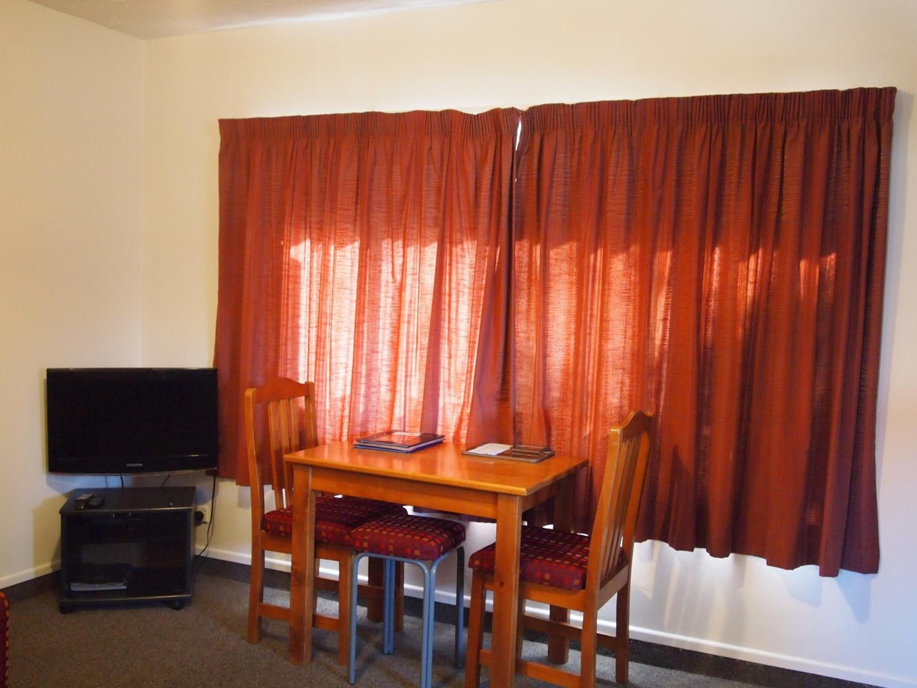 A small dining area with a table and two chairs, a television on a stand, and a window with red curtains.