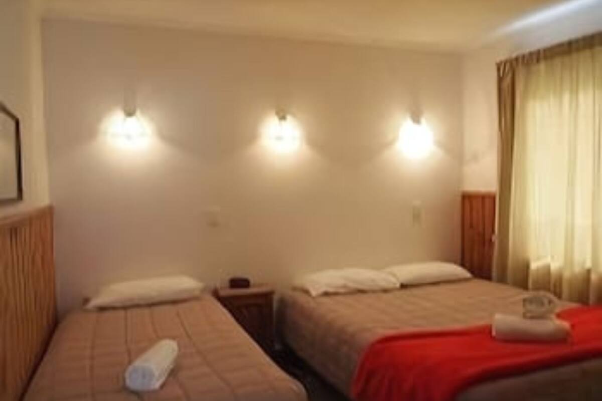 Une chambre d’hôtel avec deux lits, chacun équipé d’un oreiller et d’une serviette enroulée. On y trouve des lampes fixées au mur et une fenêtre avec des rideaux.