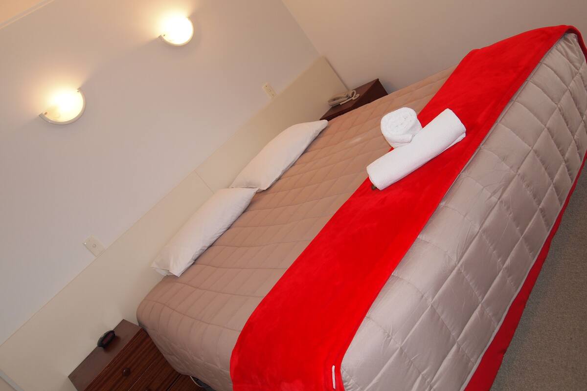 Un lit bien fait, avec une couverture rouge et deux serviettes enroulées, une table de chevet en bois et un luminaire fixé au mur.
