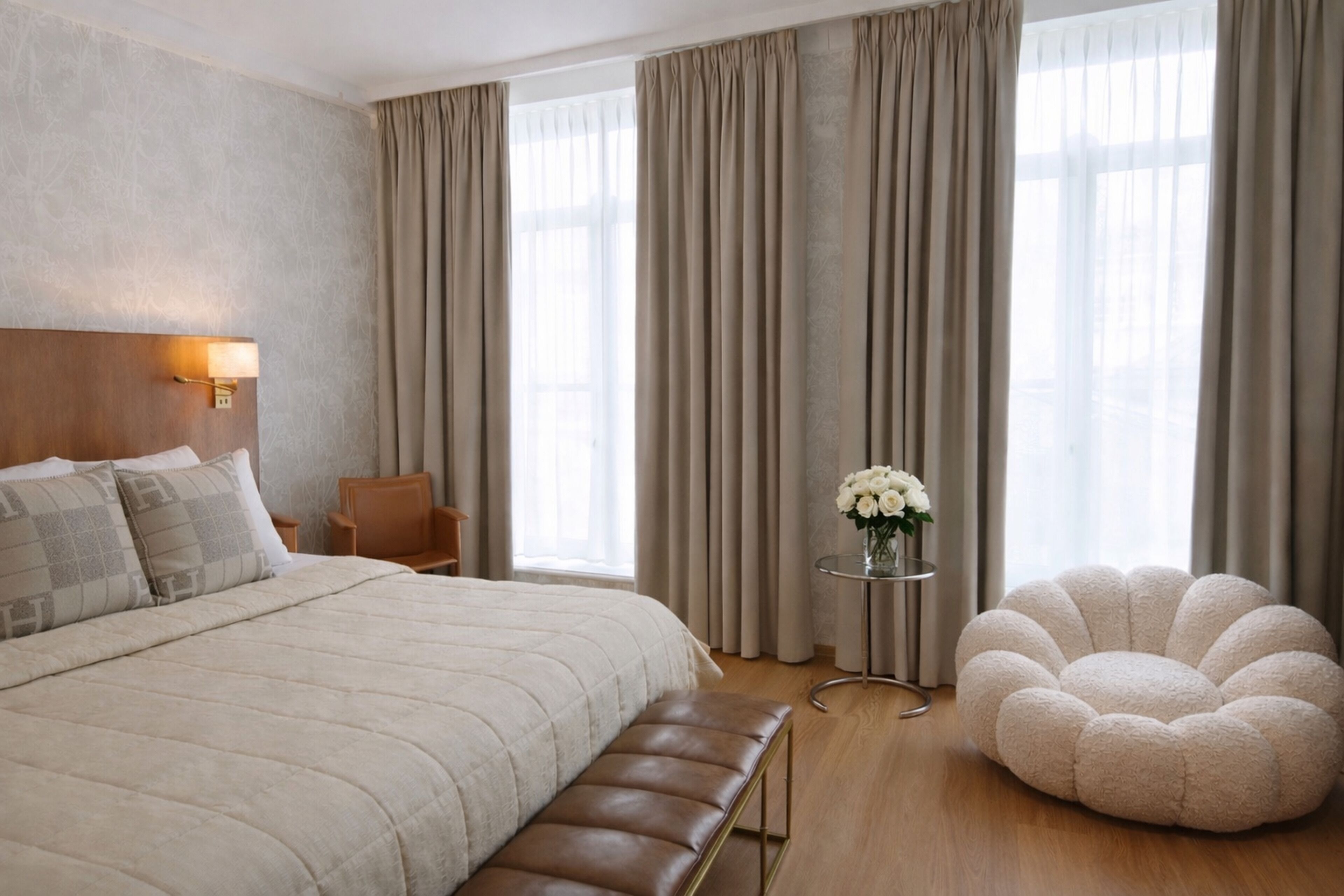 A hotel room with a large bed, a bench, a round chair, a small table with flowers, and a view through sheer curtains.