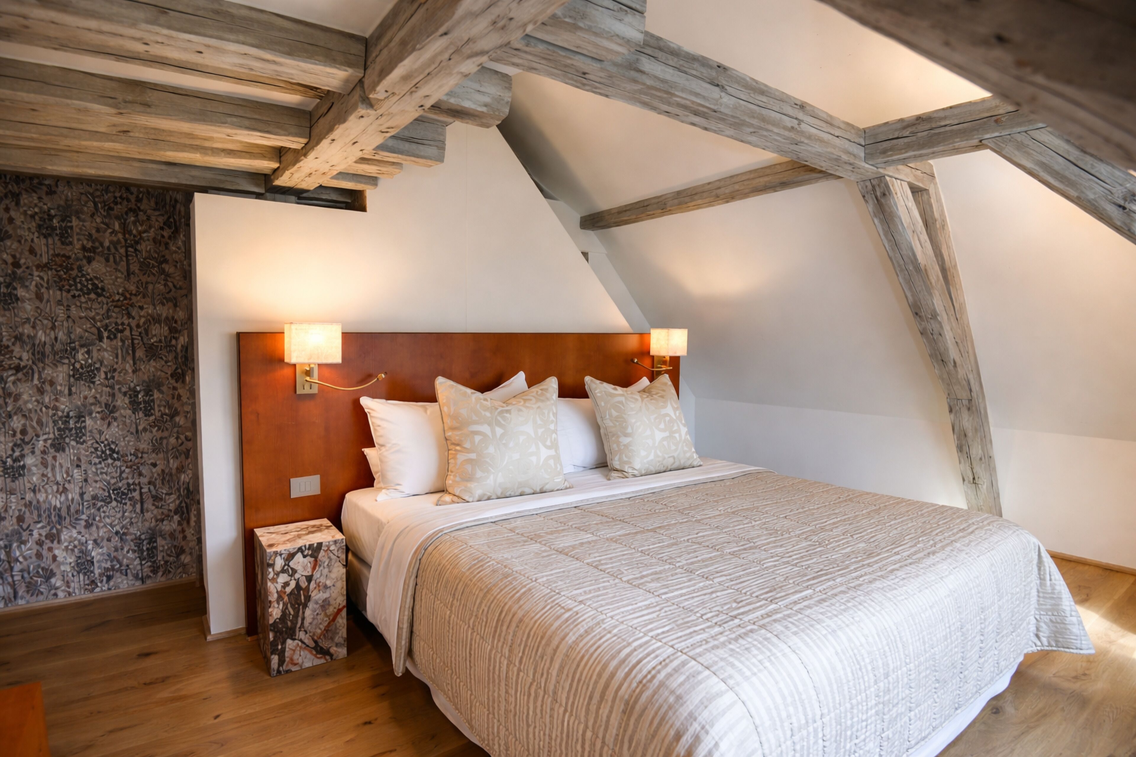 A bedroom with a sloped ceiling, wooden beams, a bed with a headboard, and two wall-mounted lamps.