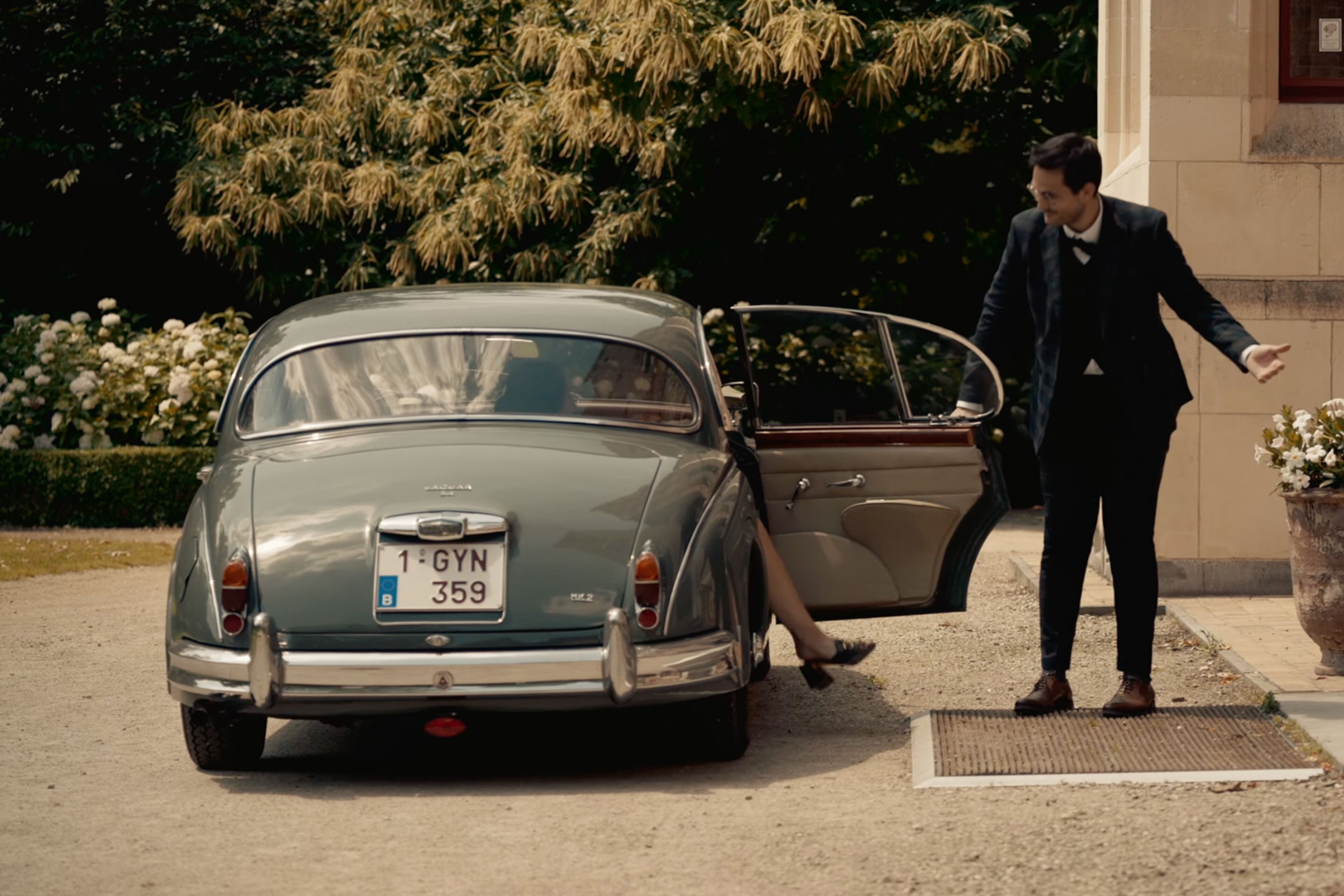 Une voiture ancienne est garée, sa portière est ouverte, une personne se tient à côté, et un bâtiment se trouve à l’arrière-plan.