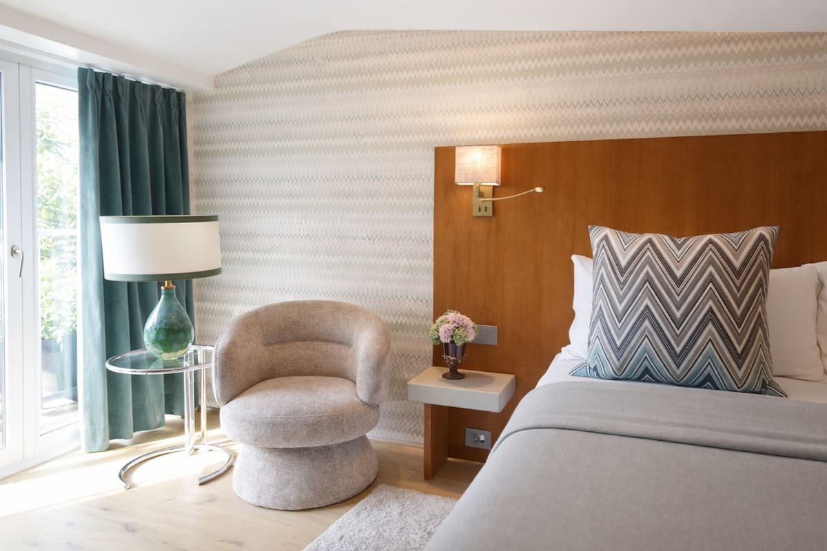 A modern hotel room with a bed, a chair, a nightstand, a lamp, and a wall-mounted light fixture.