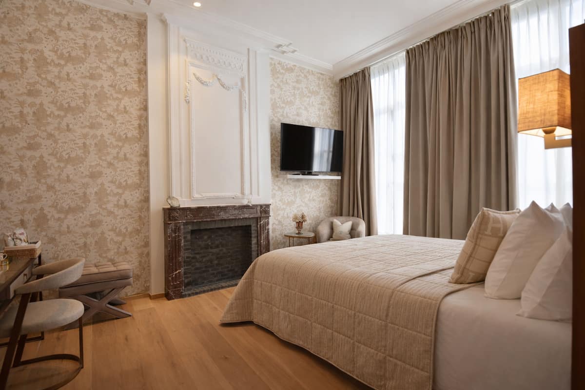 A bedroom with a large bed, a flat-screen TV mounted above a fireplace, a dining area with chairs, and a view of the outdoors through curtains.
