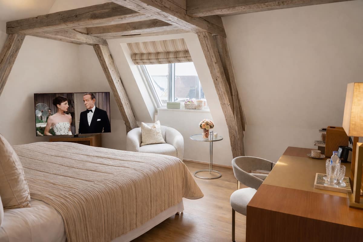 A bedroom with a bed, a TV, a chair, a small table, and a window.
